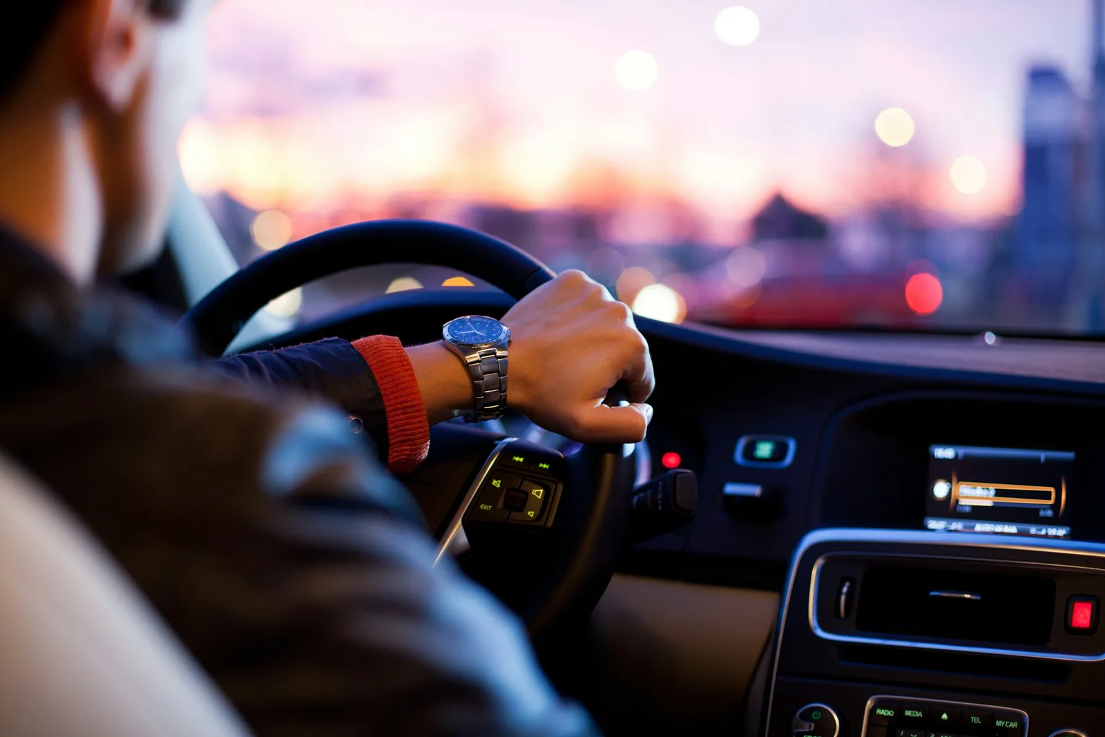 A caregiver driving a personal vehicle between client visits, hands on the steering wheel and wrist watch visible at midday