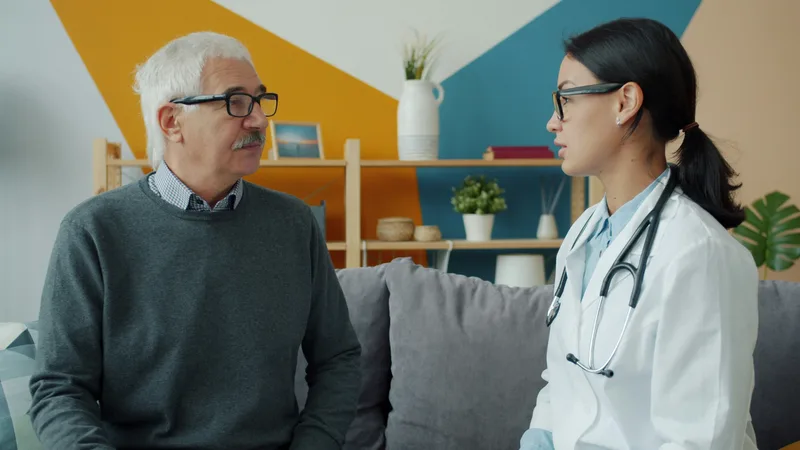 Doctor in a blue shirt seated across from an elderly patient on a couch during a home consultation visit