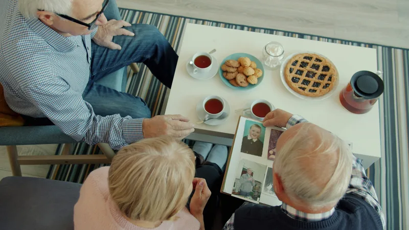 Two older adults seated at a table at home looking through a photo album together, with tea cups and snacks nearby