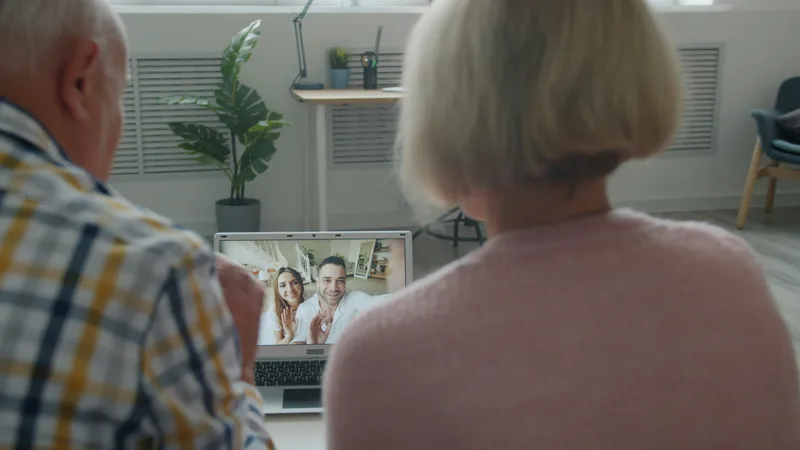 Elderly couple seated at a table waving and smiling at a laptop screen during a video call
