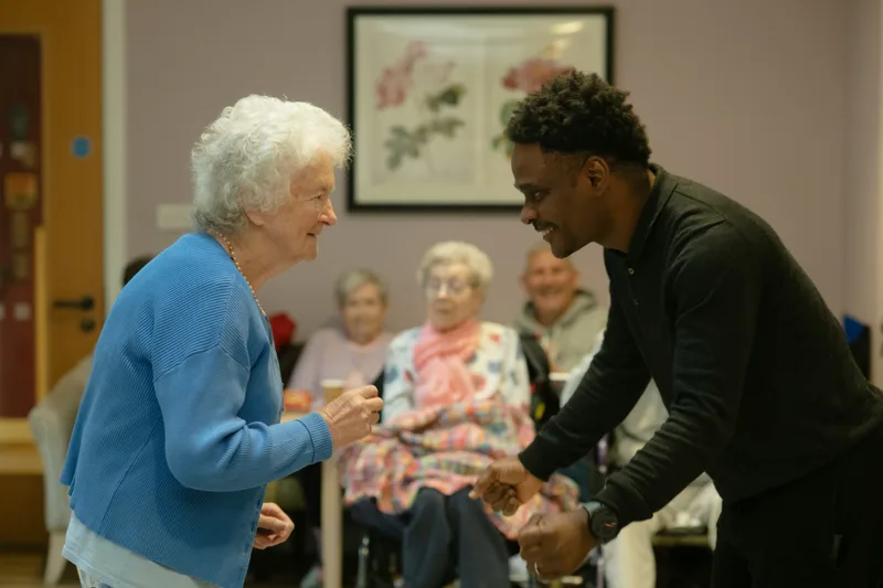 Elderly woman in a colorful outfit dancing with a younger man in a community hall setting