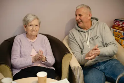 An elderly woman and an elderly man seated in chairs, both clapping and smiling in a bright community room