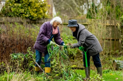 Two older women gardening side by side outdoors, both engaged and smiling in a green garden setting