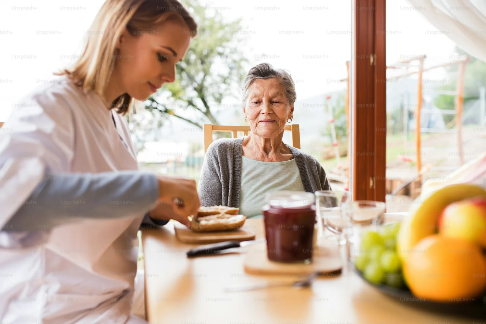 A nurse sits at a kitchen table with an elderly woman during a home visit, reviewing a plan
