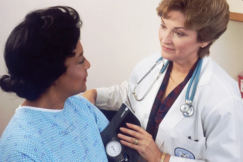 A woman with a stethoscope around her neck sitting across from another woman and speaking with her in a clinical setting