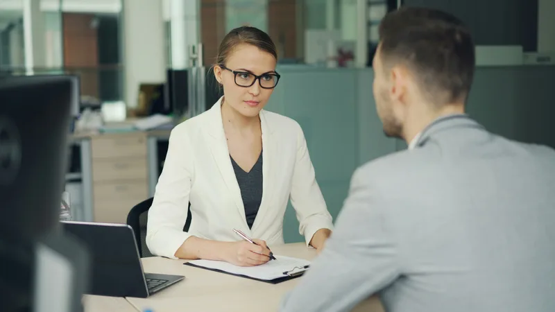 Woman in glasses conducting an interview with a man seated across an office desk, both engaged in professional conversation