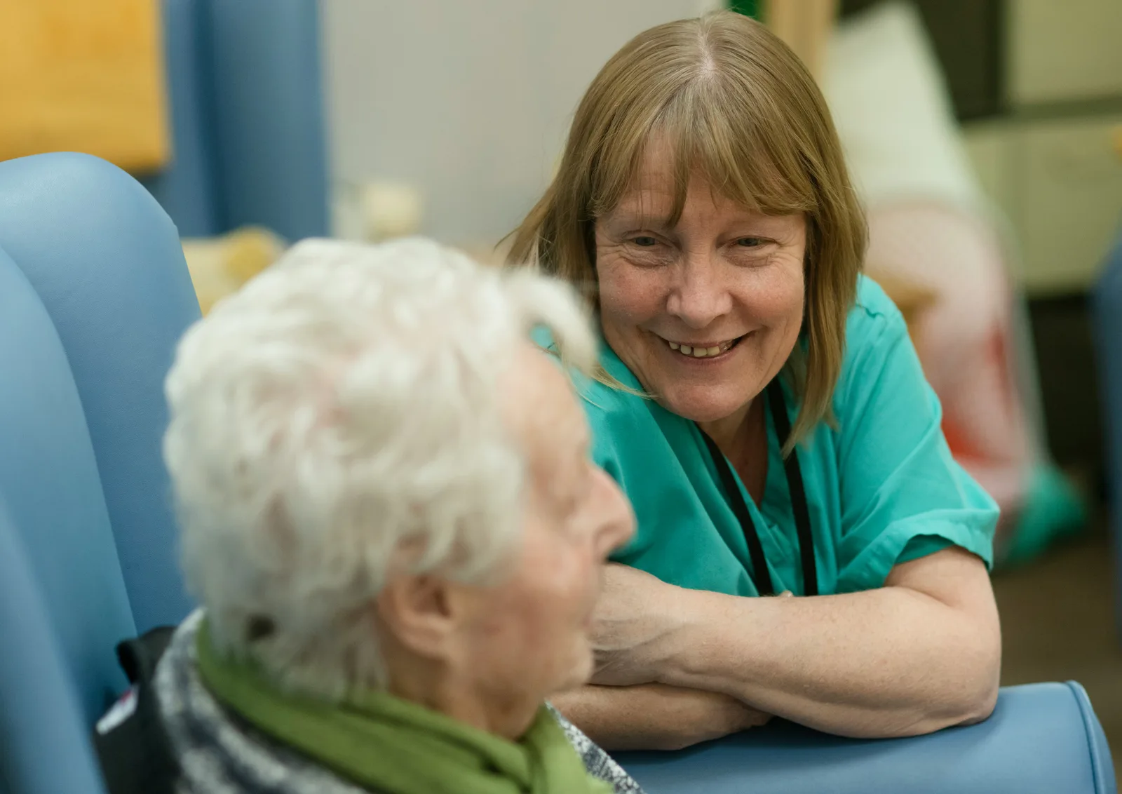 Two women seated in upholstered chairs in a home setting, engaged in conversation during a scheduled wellness check