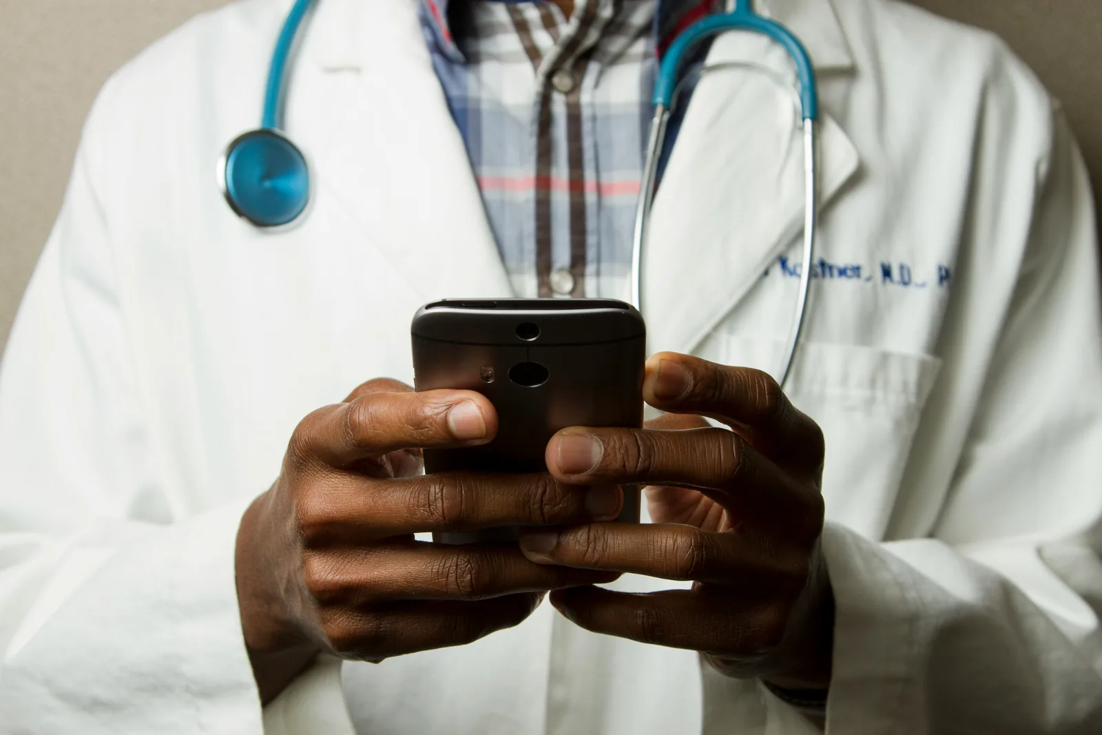 A healthcare worker in a medical gown with a green stethoscope around their neck looking at a smartphone screen while standing
