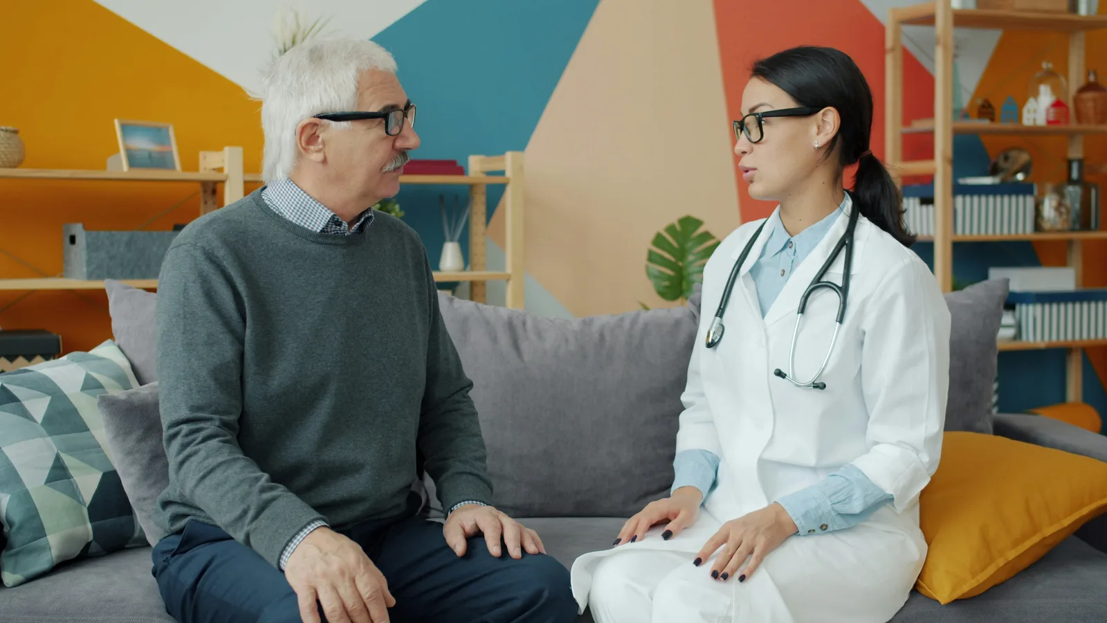 Physician sitting beside elderly male patient on a couch, reviewing paperwork during a home care supervisory visit