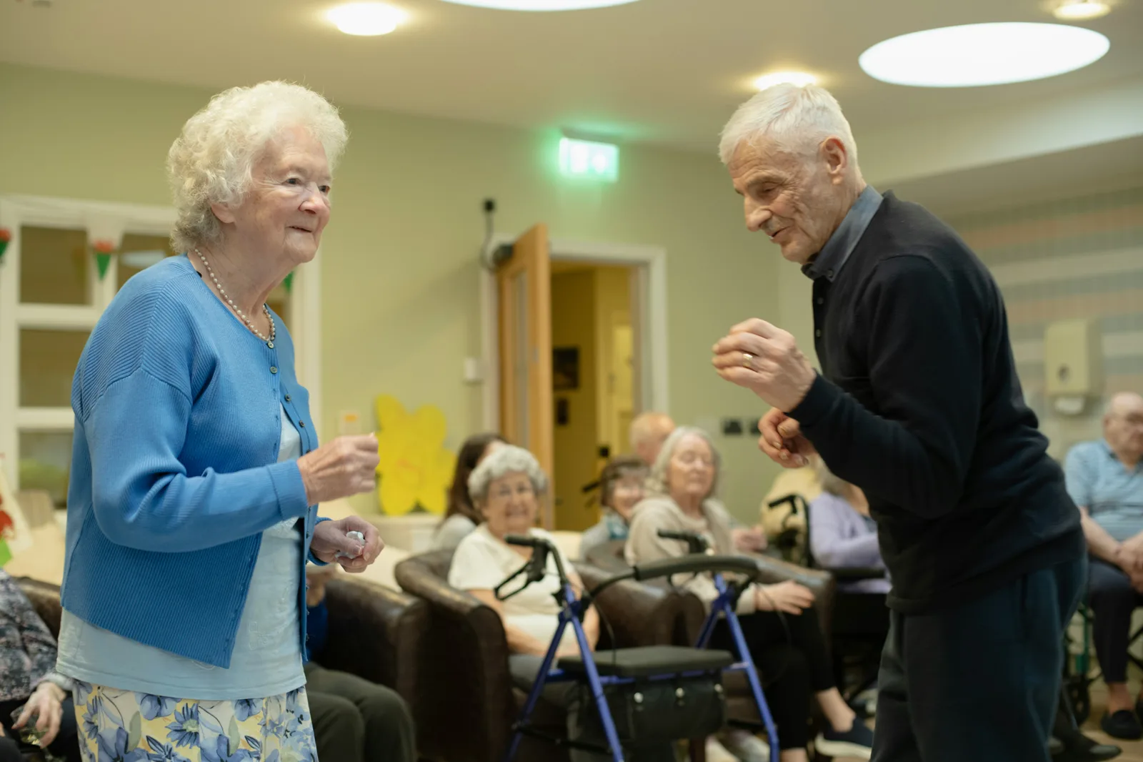 An older couple dancing together on a wooden floor while other residents watch and smile in the background