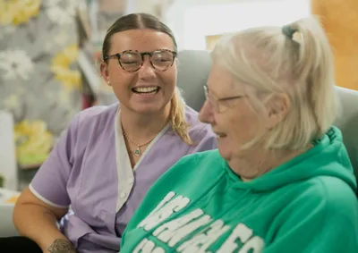 Two elderly women laughing together indoors in a warmly lit room