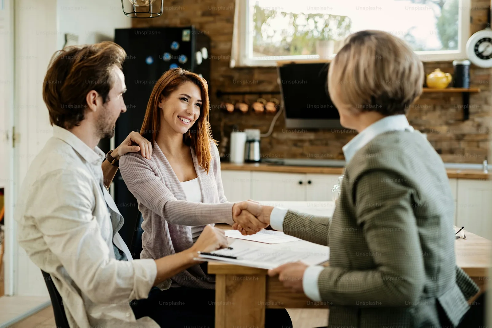 An adult couple shaking hands with a financial advisor at a kitchen table while reviewing paperwork together.