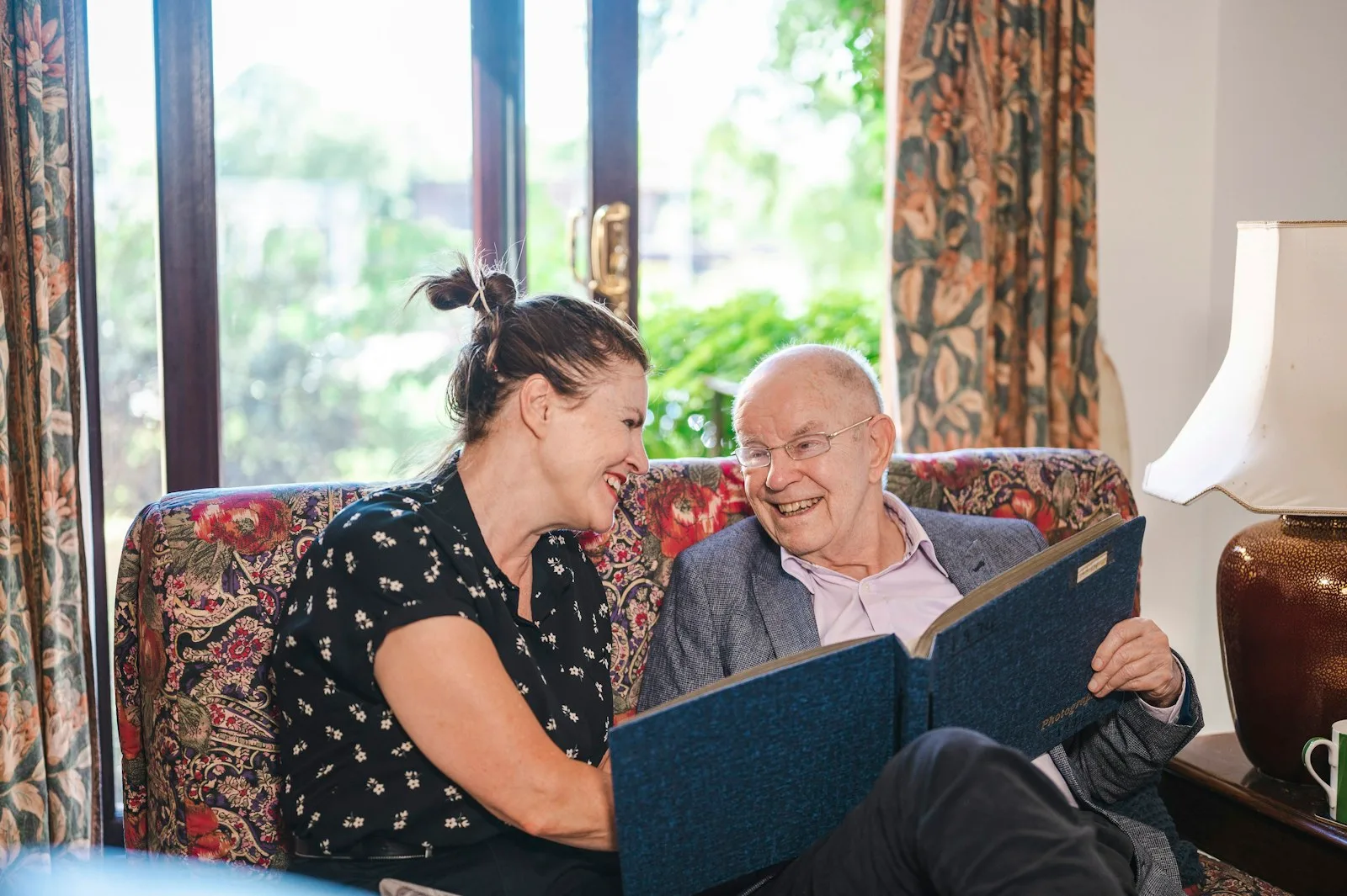 A man and a woman sitting together on a couch in a home setting, representing the personal-care visit that is the billable unit of Medicaid HCBS reimbursement.