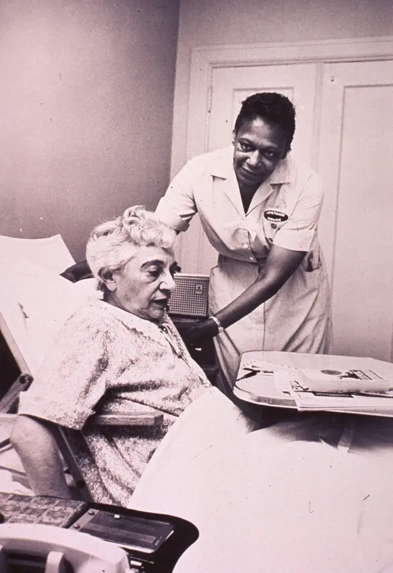 Home care aide in a patterned apron standing beside an elderly woman in a wheelchair, assisting her indoors