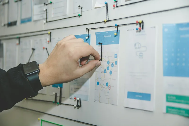 Person holding a tablet displaying a mobile app user flow, with hand-drawn wireframe workflow on paper behind it