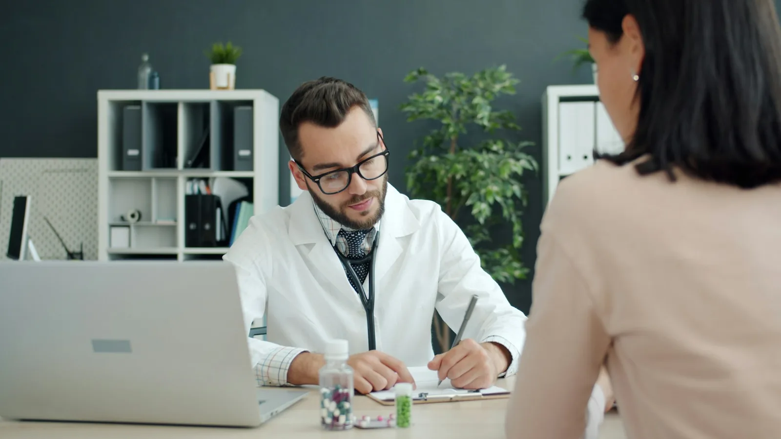 Healthcare professional writing notes on a clipboard while a patient sits across the desk