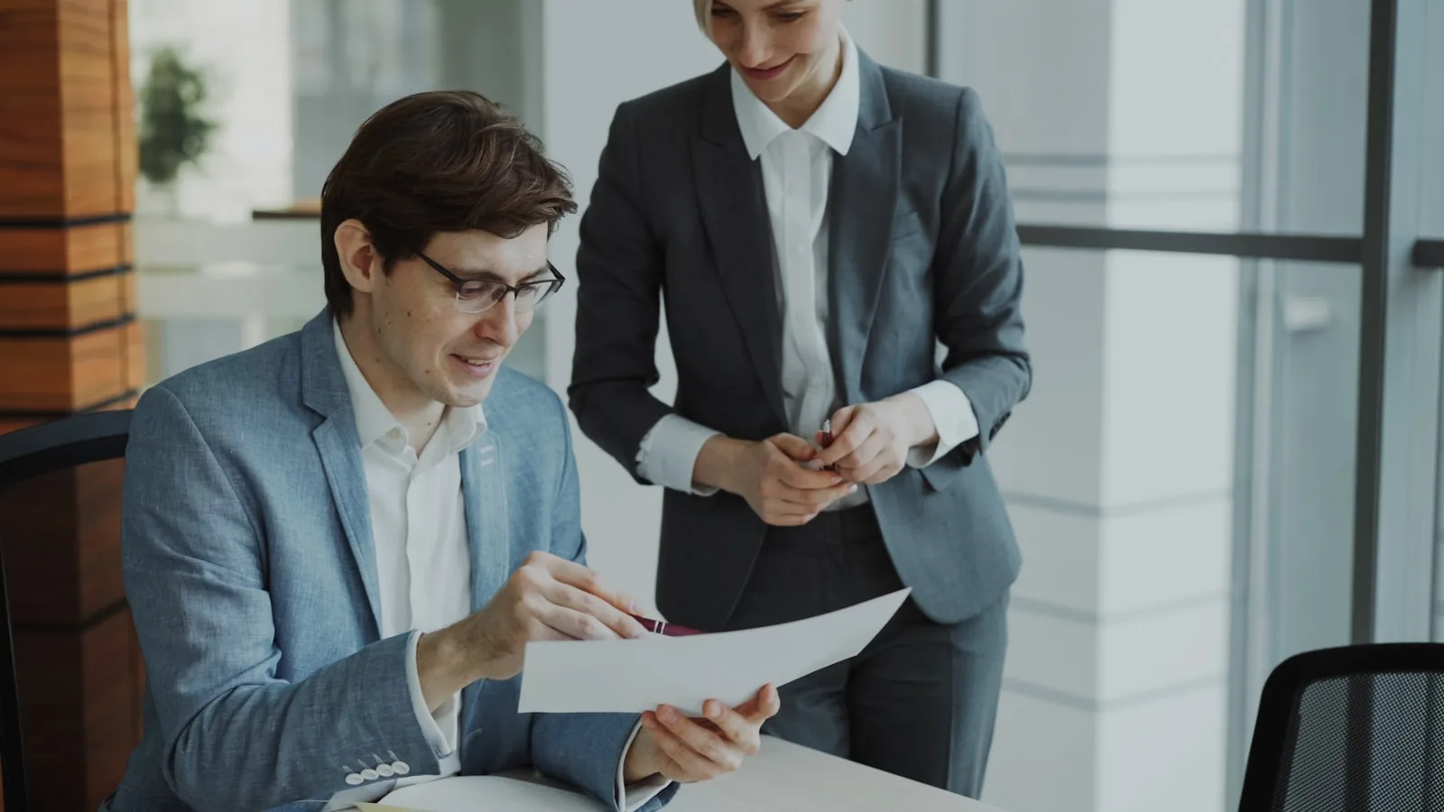 Two business professionals seated at a table reviewing printed documents and a laptop screen together.
