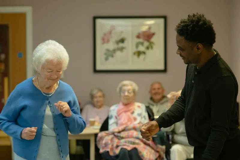 Elderly woman dancing joyfully with a younger male caregiver in a bright indoor room