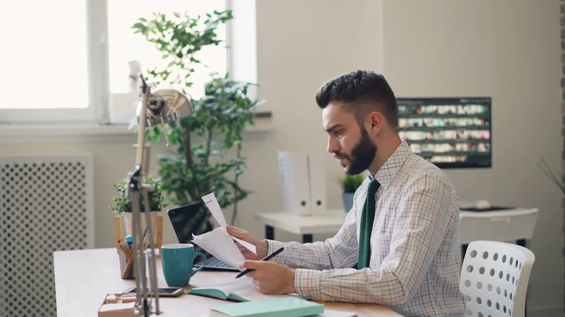 A man in a dress shirt seated at a wooden desk reviewing printed documents alongside an open laptop
