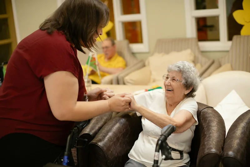 A caregiver in a blue shirt steadies an elderly woman using a metal walker as they move through a home hallway