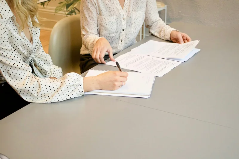 Two women seated at a desk, one signing printed documents while the other watches and prepares to countersign