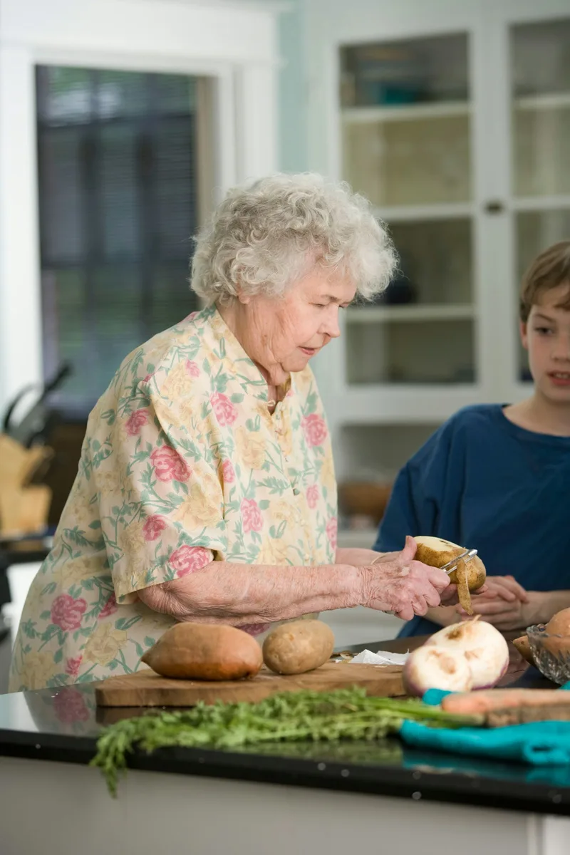 A woman in a pink floral shirt receives a wooden meal tray from a home health aide during an in-home visit
