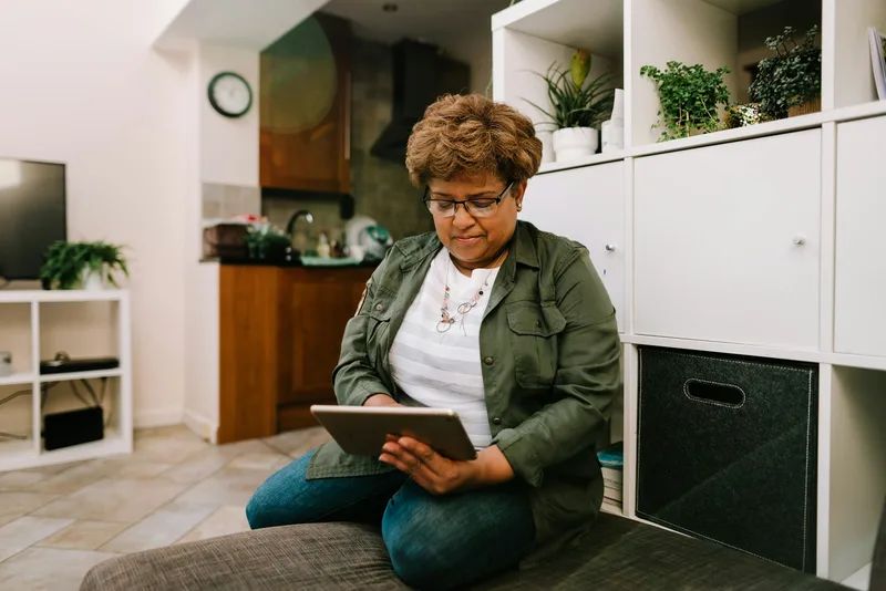 An older woman seated on the floor leaning against a grey couch, reviewing documents on a tablet computer